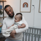 Father holding baby wearing breathing wear sleeping bag. Nanit pro camera and floor stand in the background in front of crib